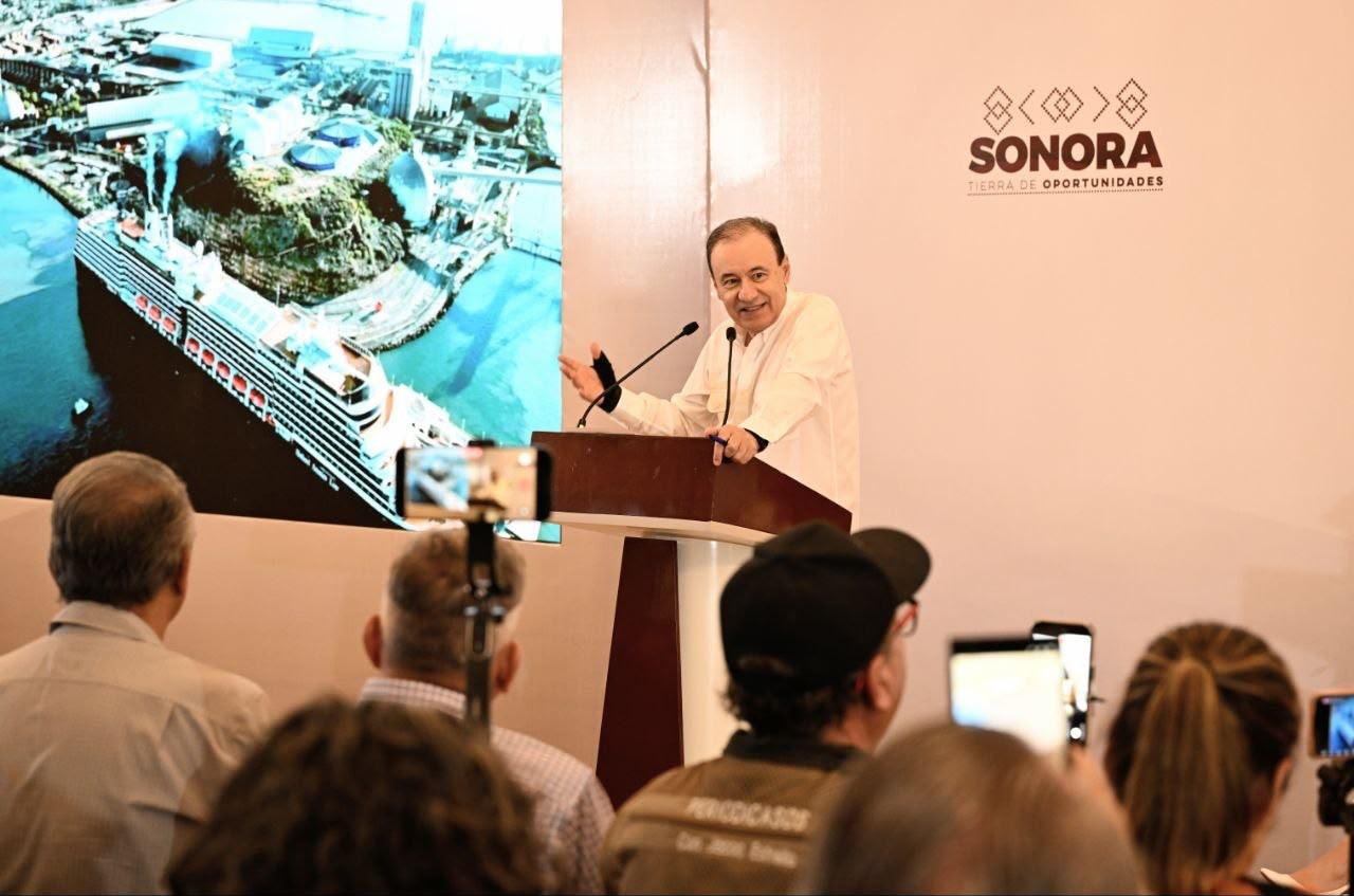 El gobernador Alfonso Durazo habla desde un pódium ante medios de comunicación, junto a una pantalla que muestra un crucero atracado