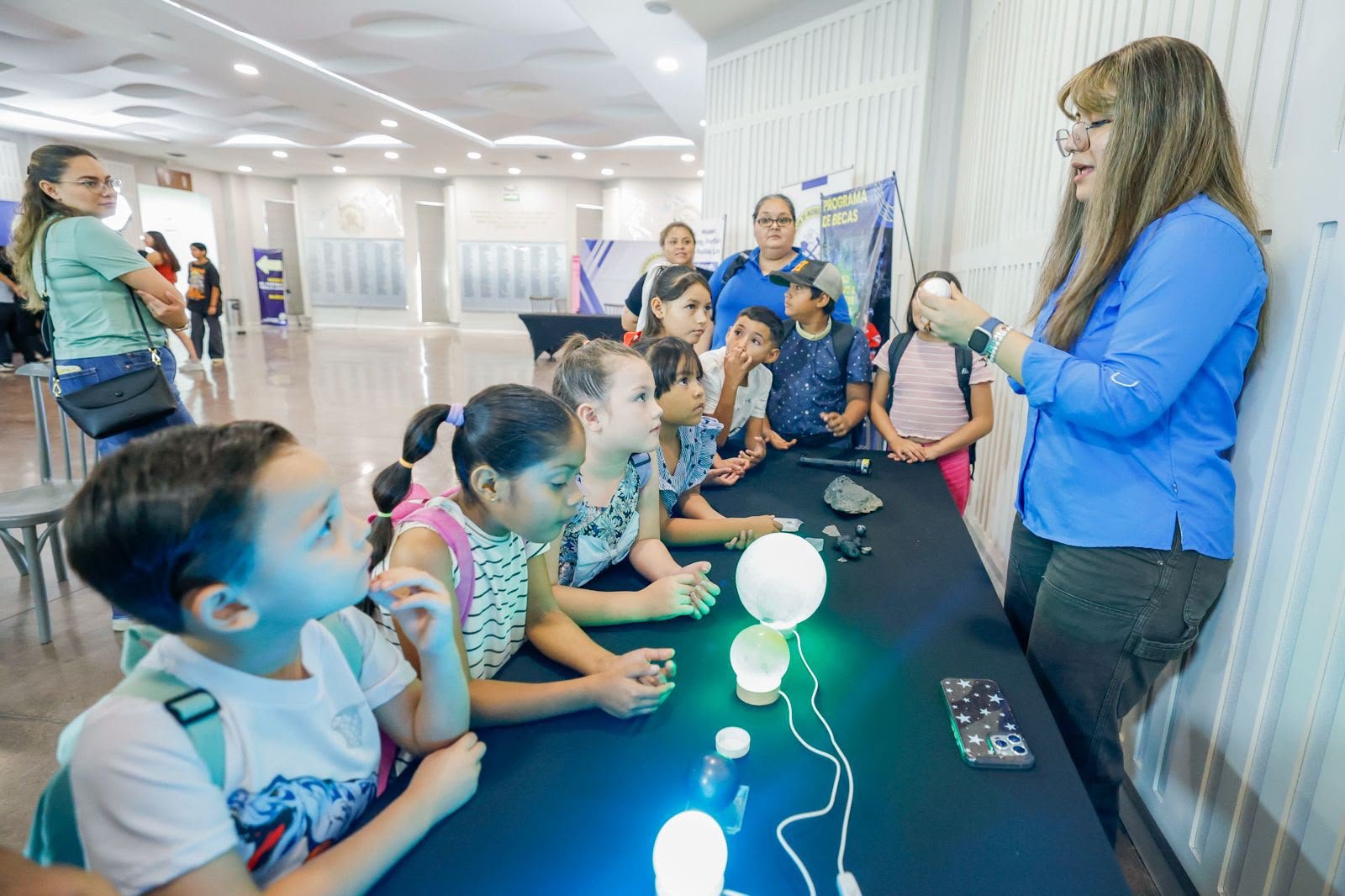 Una guía muestra minerales luminiscentes a un grupo de niñas y niños sentados frente a una mesa