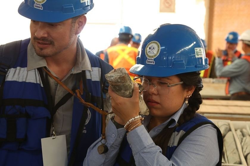 Dos personas con casco de la AIMMGM Distrito Sonora observan con lupa una muestra de mineral