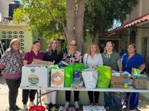 Siete mujeres de pie detrás de una mesa con cajas y bolsas de víveres donados.