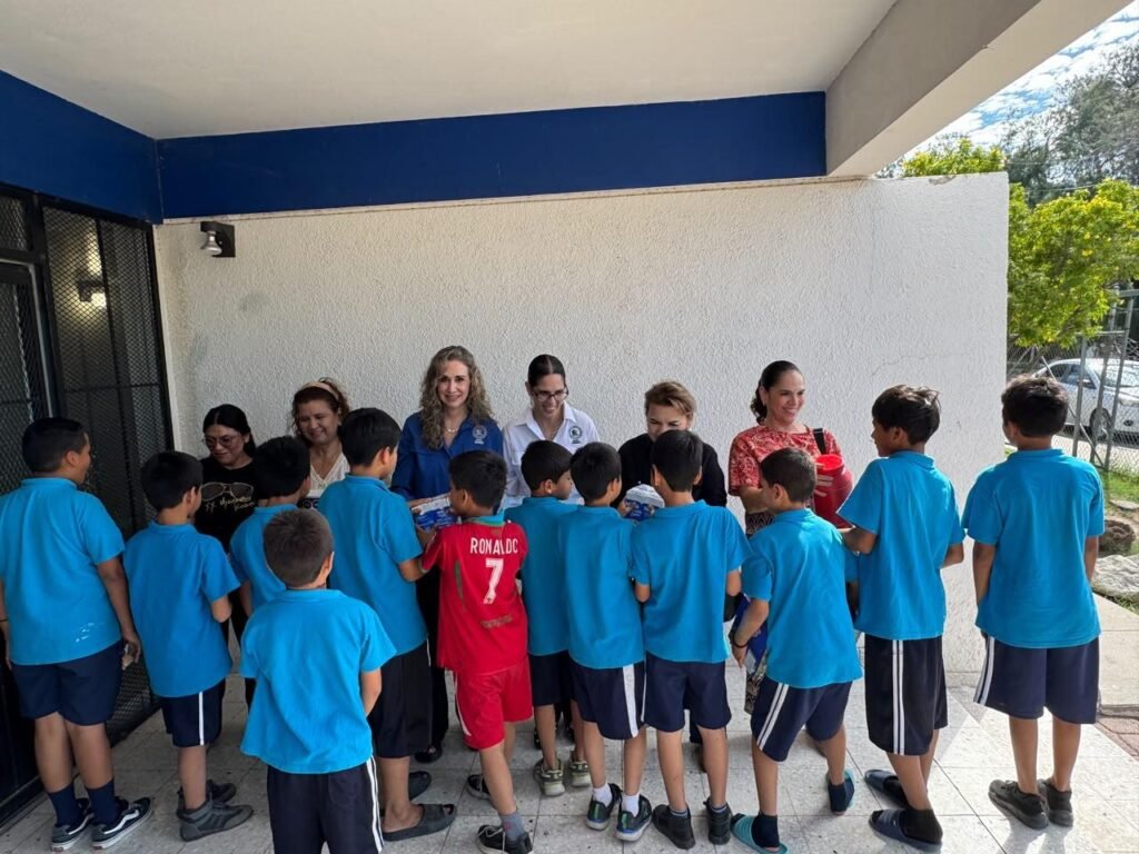 Niños con uniforme azul, la mayoría de espaldas, reciben artículos de manos de integrantes del comité.