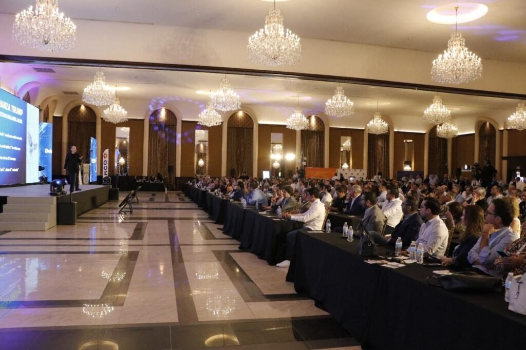 Vista lateral del salón con cientos de asistentes sentados en mesas largas frente al escenario, bajo candelabros de cristal.