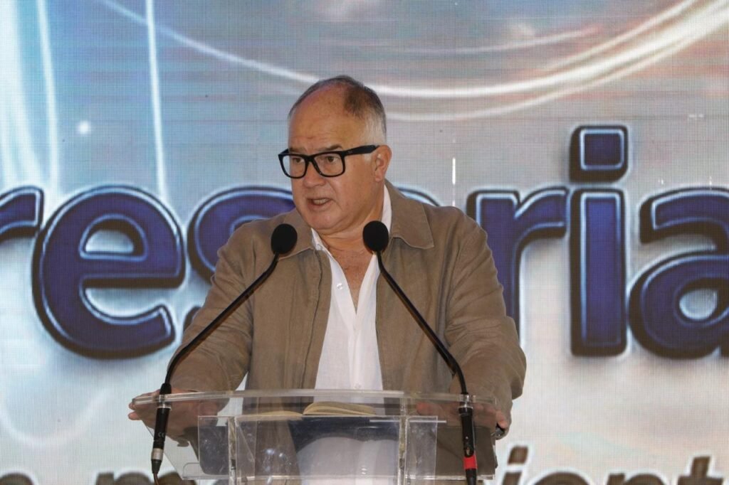 Hombre con lentes y saco beige habla en un atril transparente.
