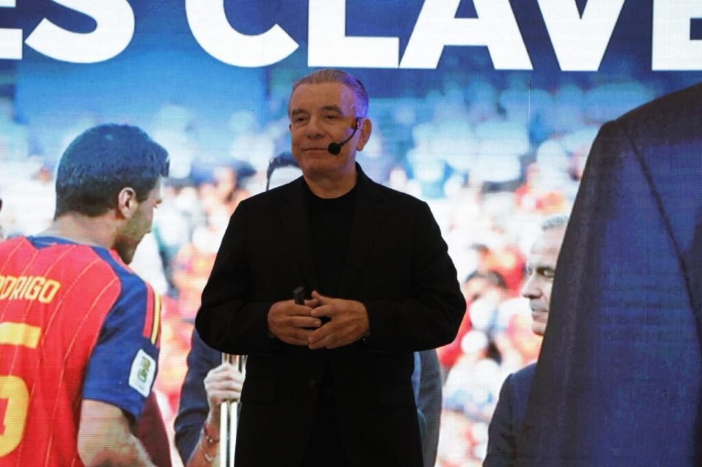 Hombre vestido de negro con micrófono de diadema frente a una pantalla con una imagen de futbol.