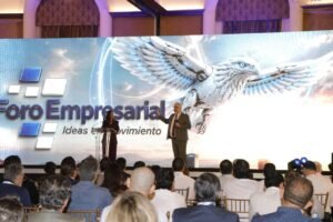 Escenario del Foro Empresarial Ideas en Movimiento con una pantalla gigante de un águila robótica; una mujer en el atril y un hombre habla frente al público.