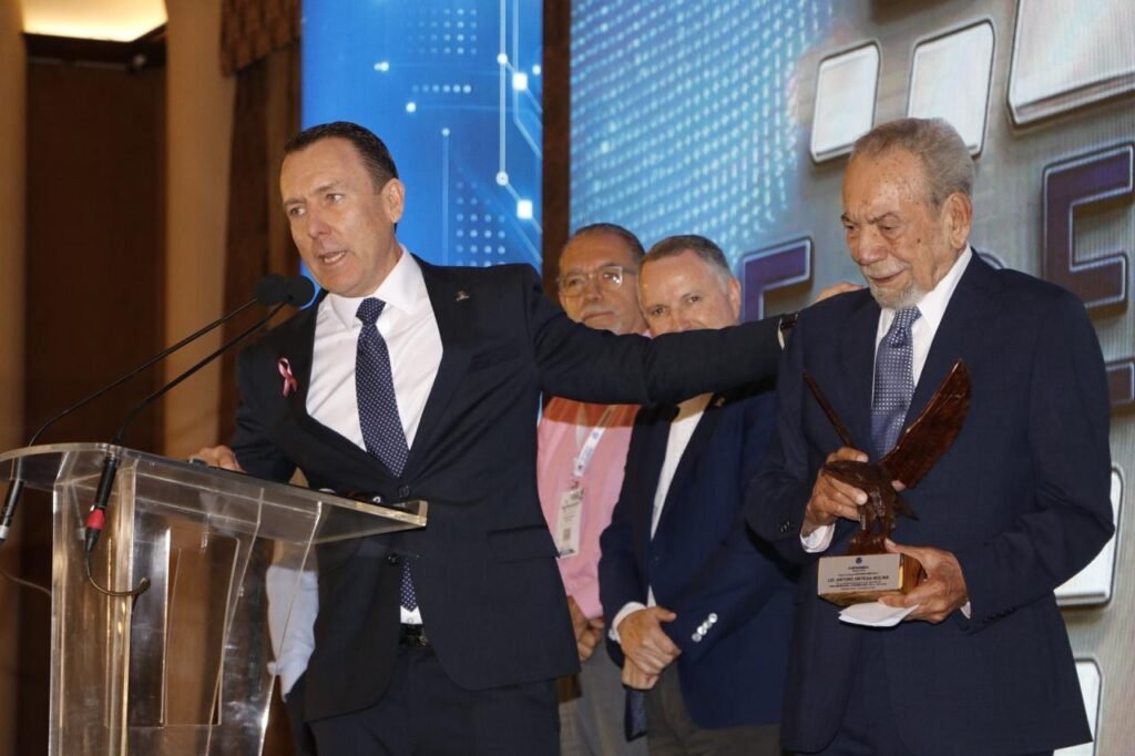 Un hombre habla en el atril mientras abraza a un hombre mayor que sostiene un trofeo con forma de águila; la placa dice «Lic. Arturo Ortega Molina».
