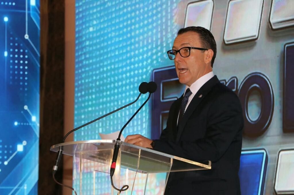 Hombre de traje oscuro y lentes habla en un atril transparente con dos micrófonos.