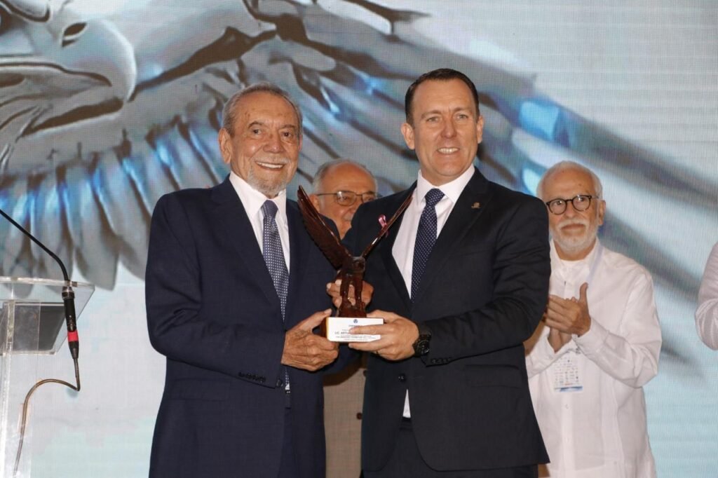 Dos hombres de traje sostienen juntos un trofeo con forma de águila sobre el escenario.