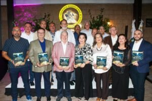 Grupo de personas de pie sostiene ejemplares del libro “La Veta Humana” frente al escudo de la AIMMGM Distrito Sonora.