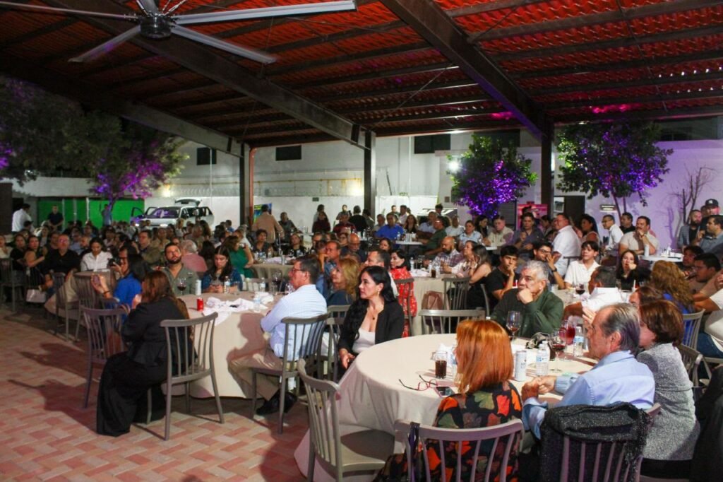 Vista general de los asistentes sentados en mesas redondas bajo una techumbre.
