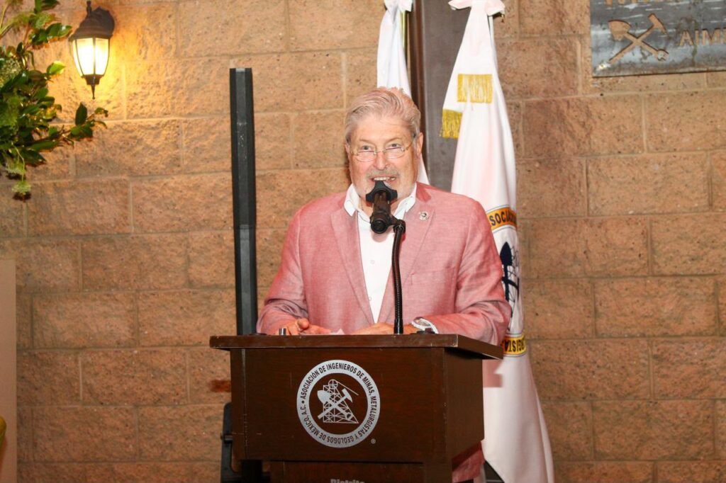 Hombre con saco rosa habla desde el atril con el escudo de la AIMMGM.