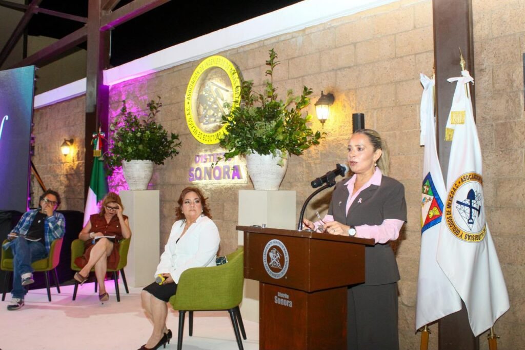Mujer con blusa rosa habla desde el atril; al fondo, tres personas sentadas en el escenario.