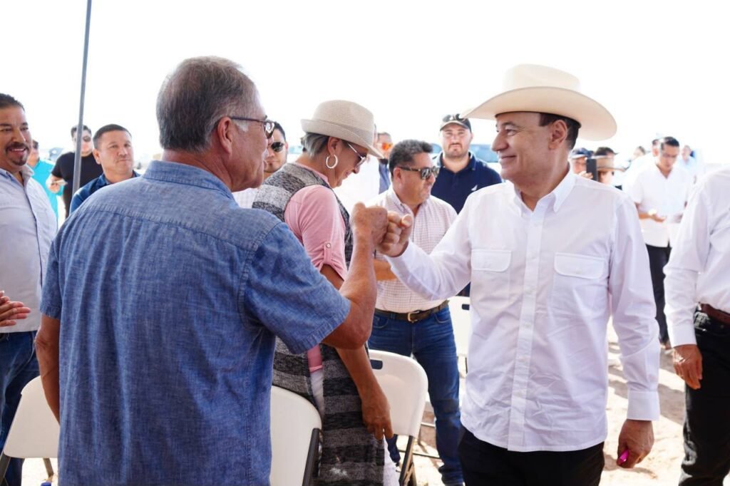 Hombre con sombrero y camisa blanca saluda chocando el puño con un hombre de camisa azul, rodeados de asistentes.