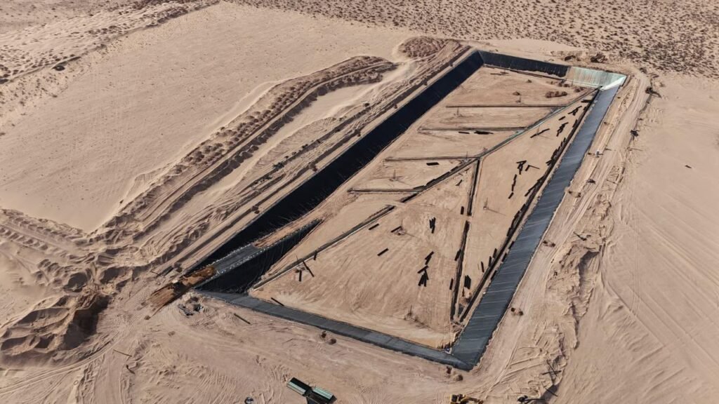 Vista aérea de una celda rectangular recubierta con geomembrana negra, rodeada de dunas de arena.
